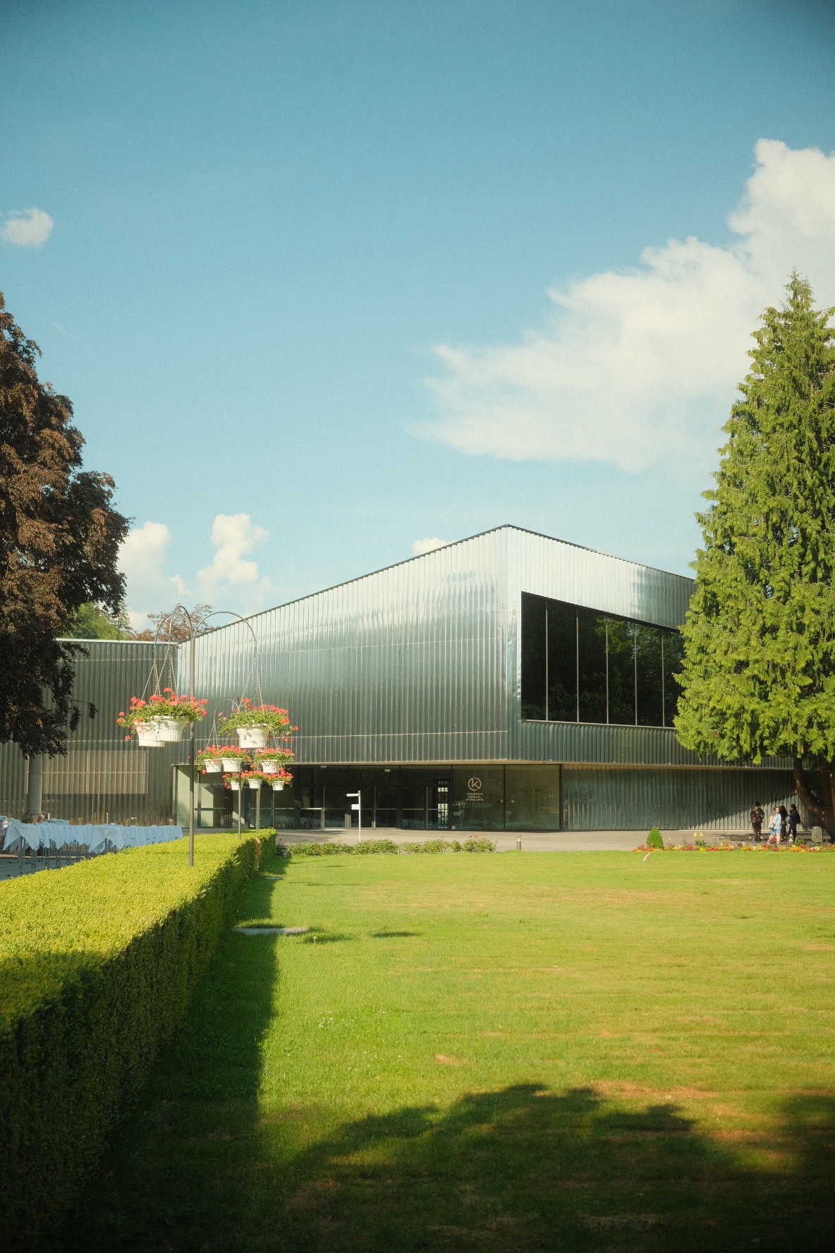 Kongress Kursaal Interlaken auditorium clad in shimmering metal, framed by tall firs and a manicured lawn.