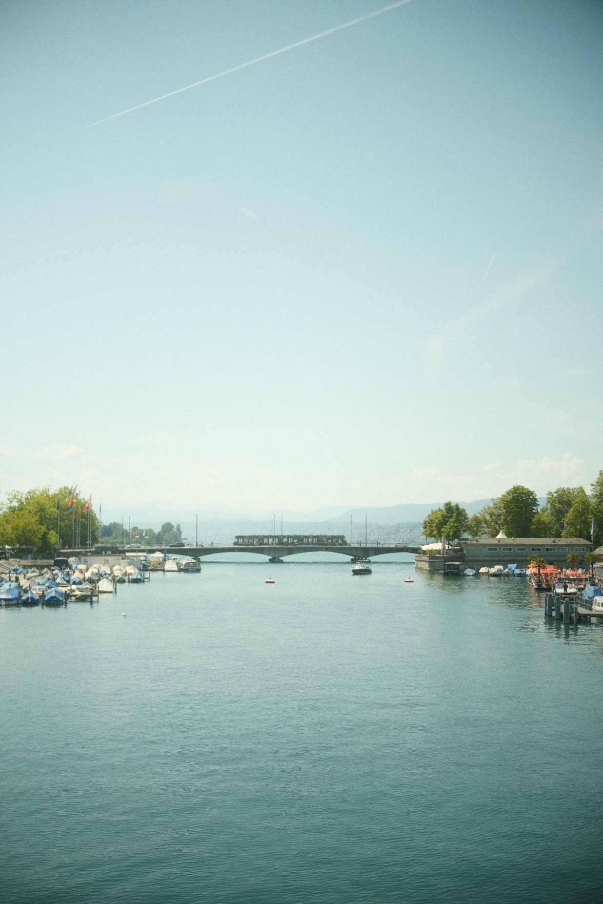Boats moored along Lake Zurich as a tram crosses the Quaibrucke under a hazy summer sky.