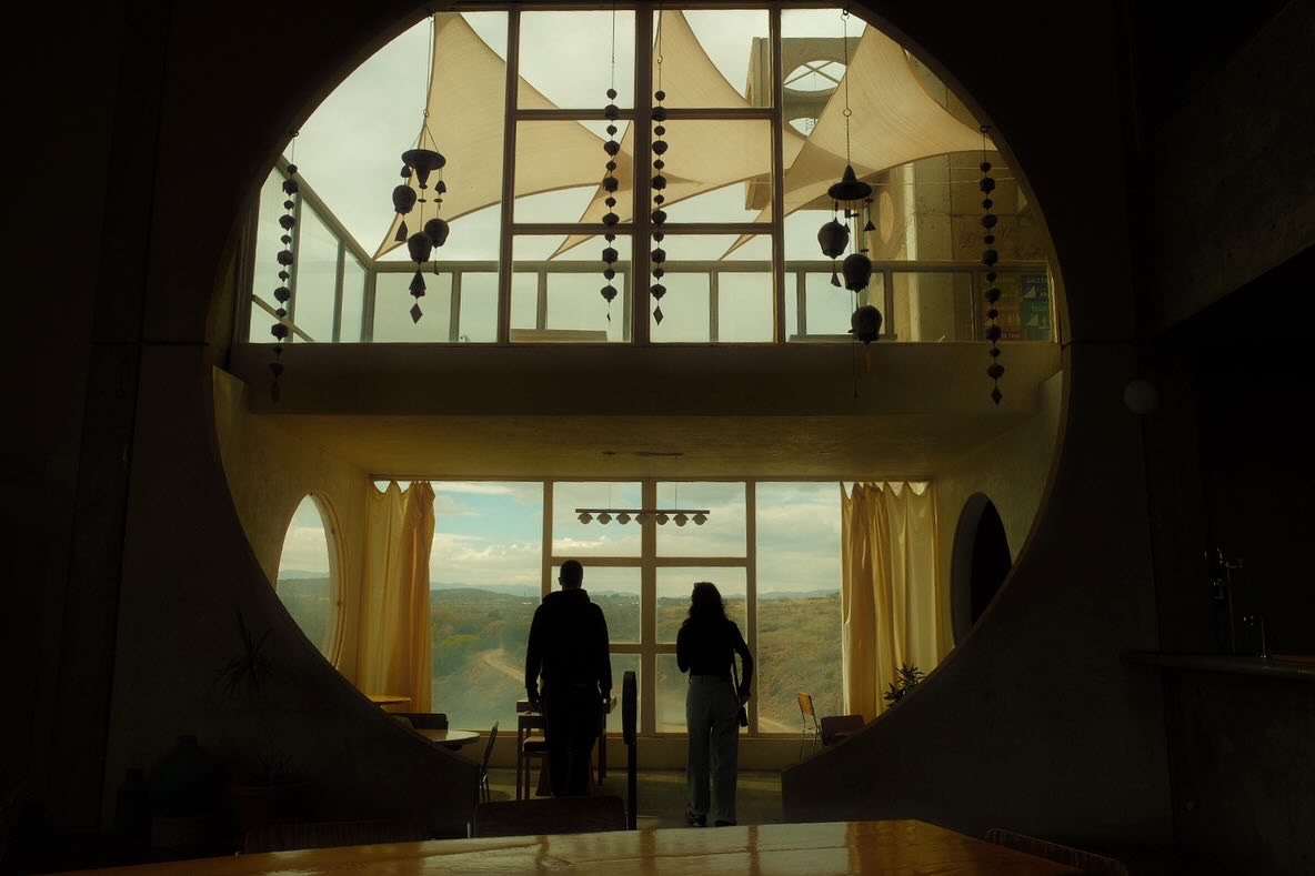Two people silhouetted against the apse window of Arcosanti, ceramic wind bells hanging in the desert light, Arizona.