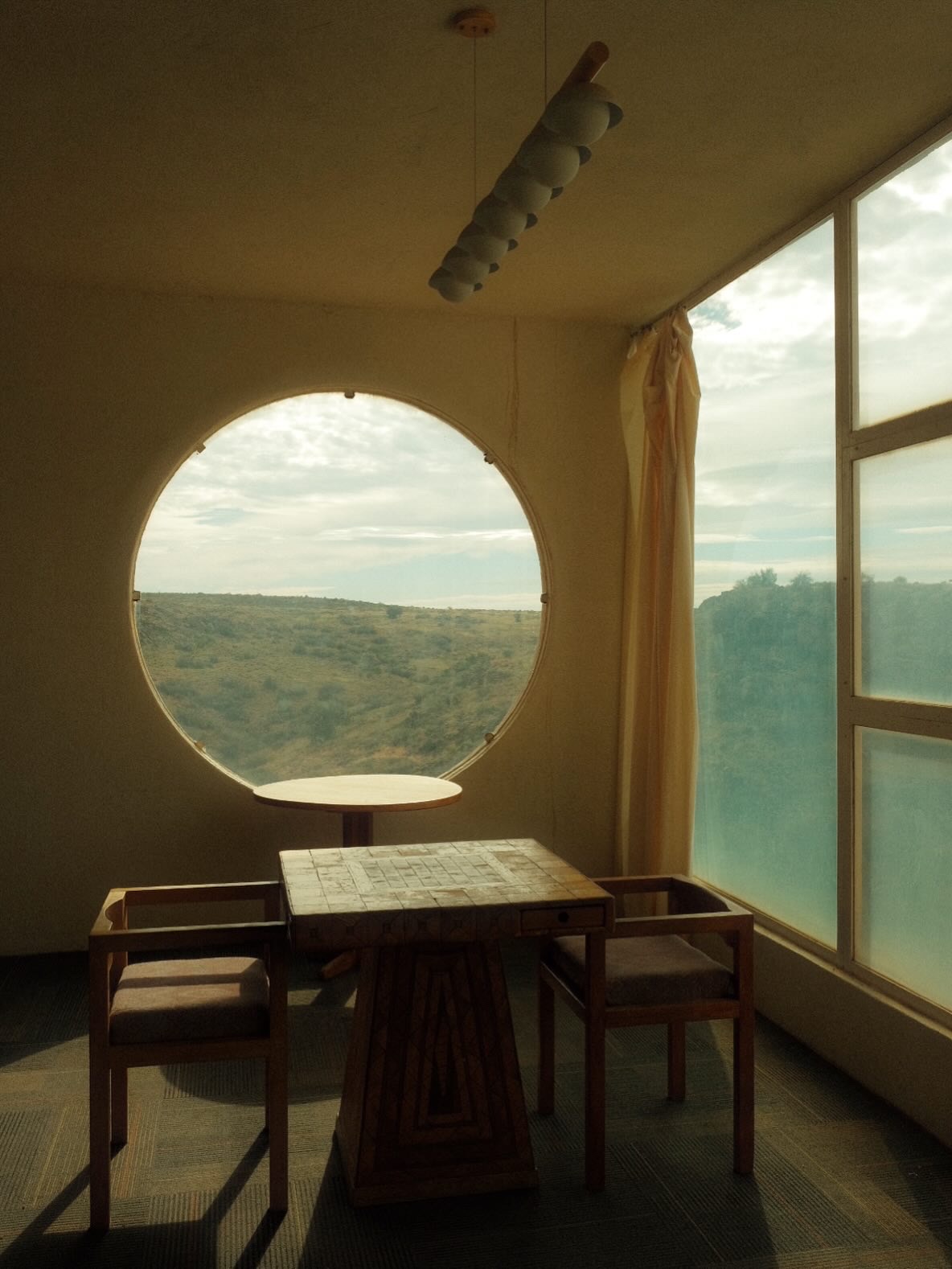 Wooden table and chairs beside a round oculus window framing the open desert at Arcosanti, Arizona.