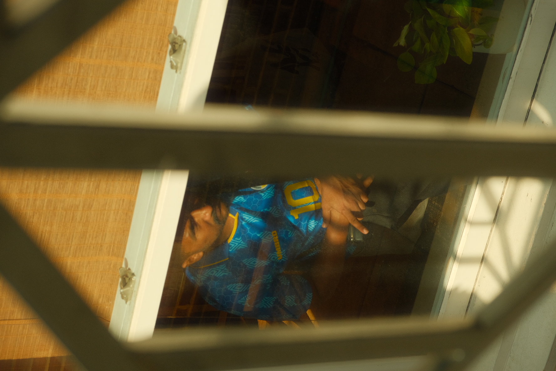Man in a blue number ten soccer jersey glimpsed through a bamboo-shaded café window in warm afternoon light, New York.