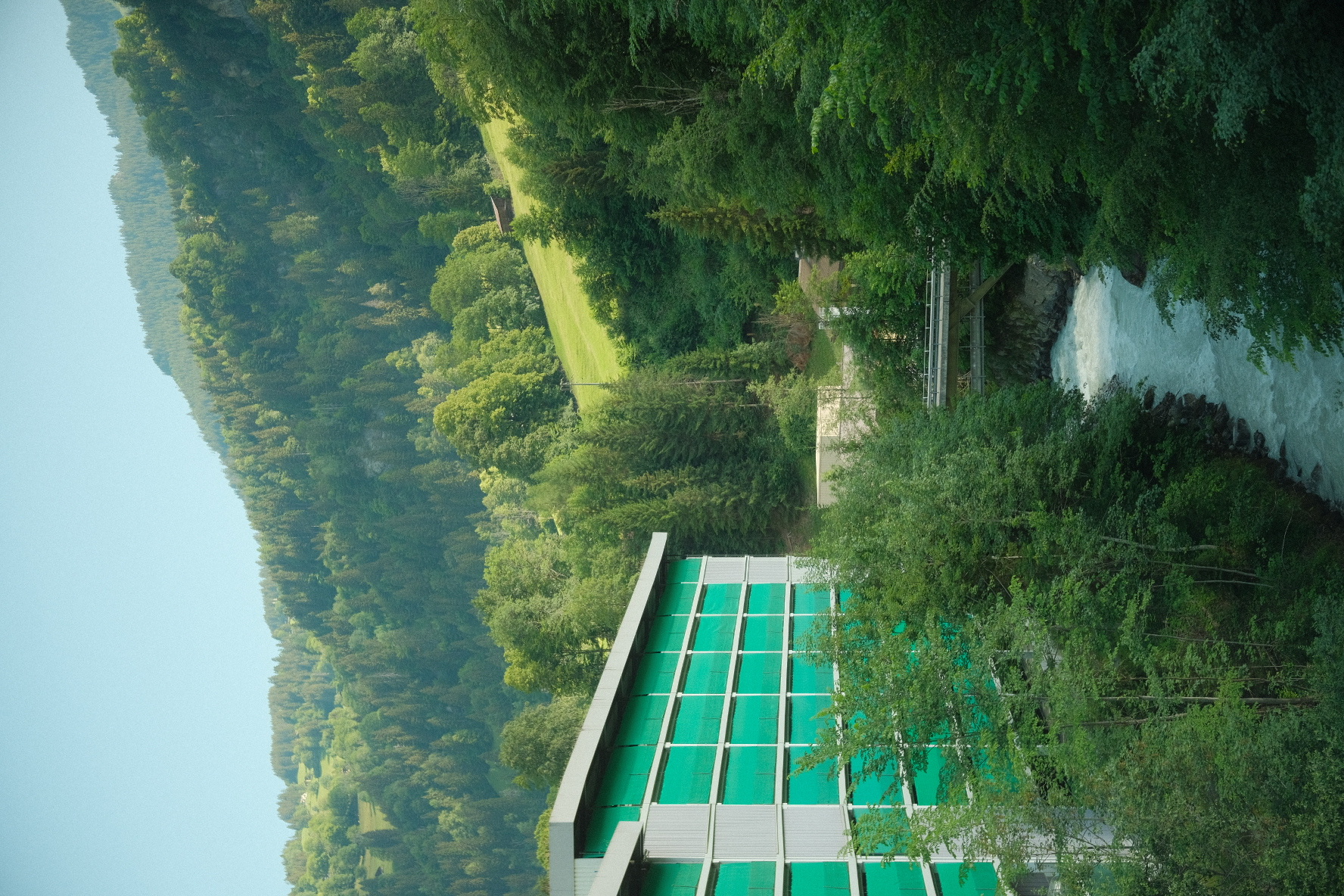 Turquoise-glassed modern building tucked into a forested riverbank beside pale glacial water.