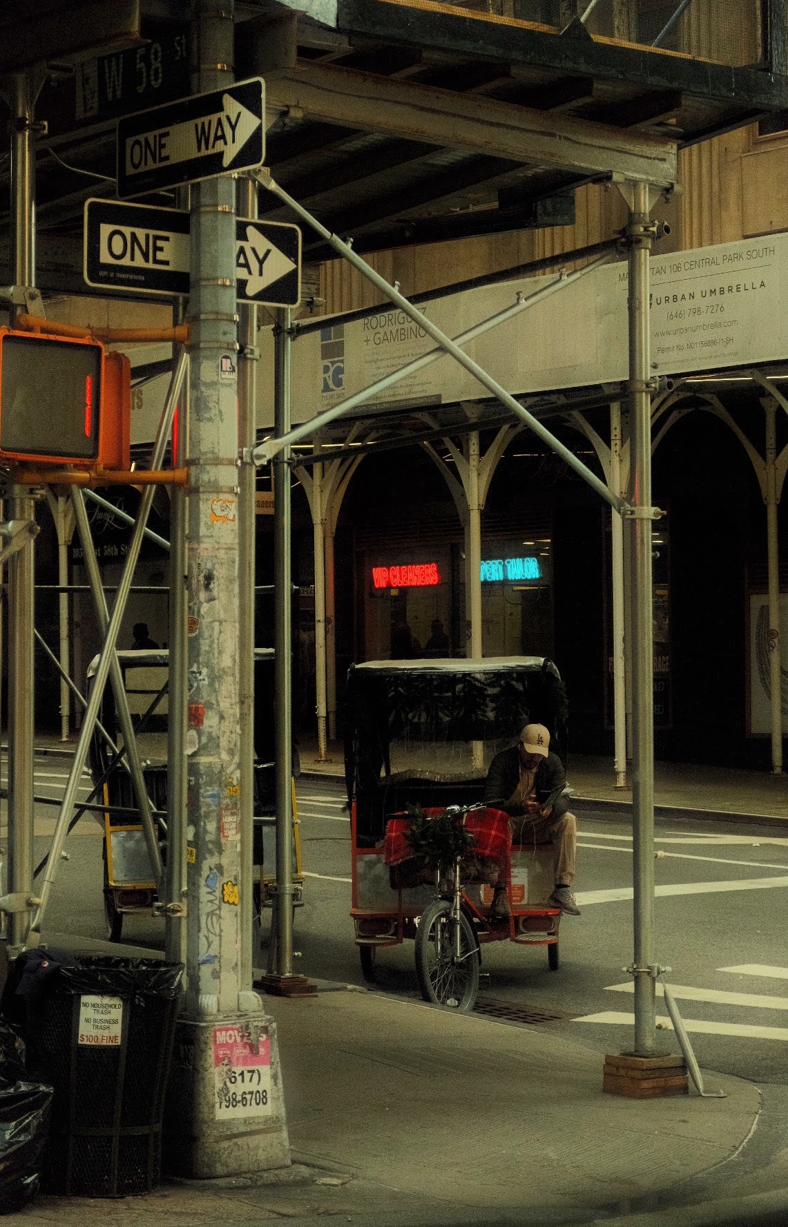 Pedicab driver resting under scaffolding at West 58th and Central Park South, one-way signs and neon cleaners sign in the background, New York.
