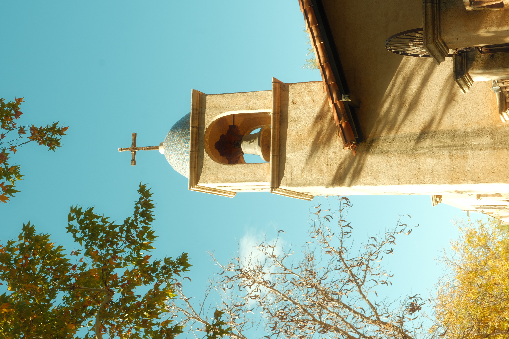 Adobe bell tower with a tiled mosaic dome and iron cross against a blue sky, framed by autumn trees in Arizona.