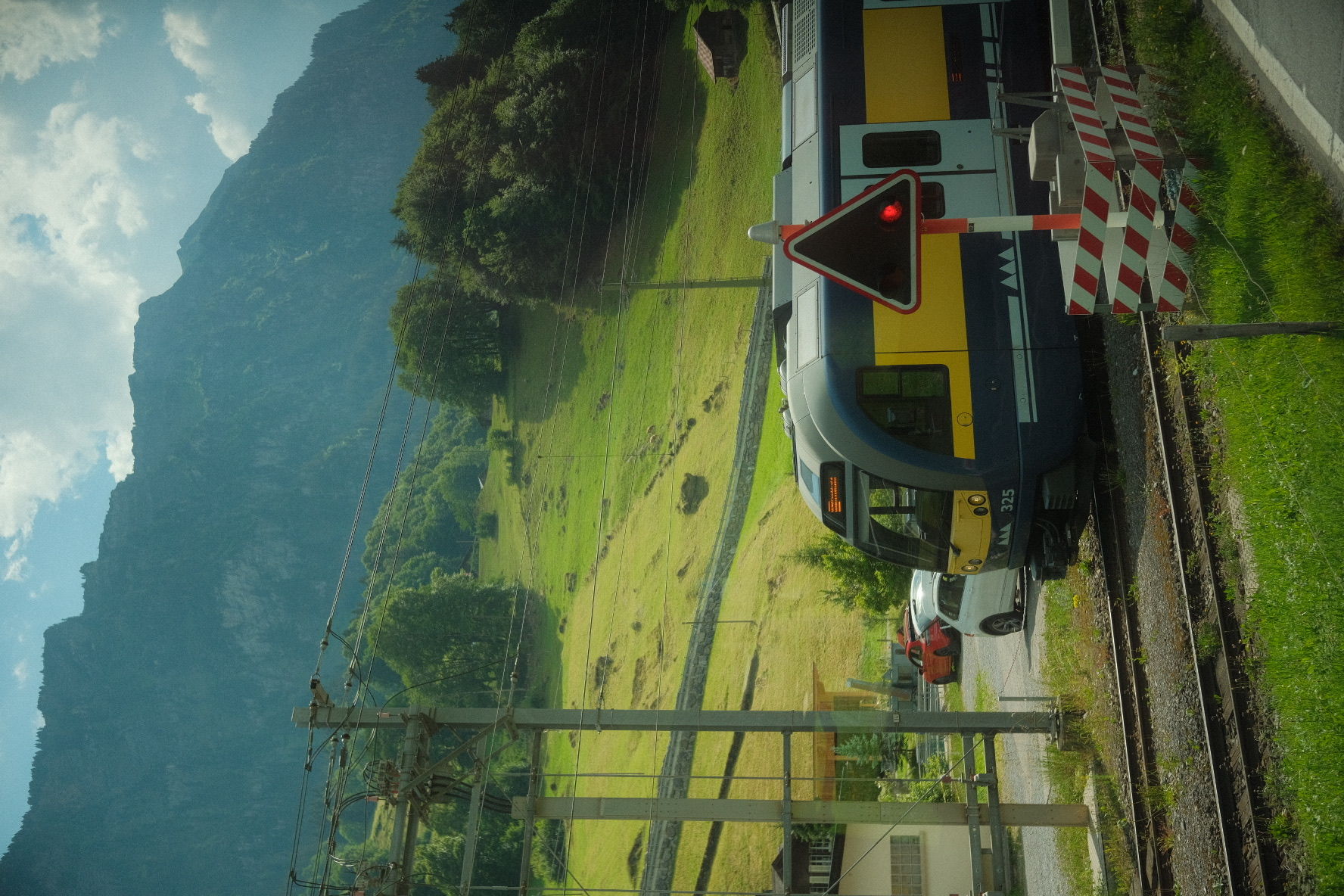 Blue-and-yellow Swiss regional train at a level crossing with a green valley and steep mountains beyond.