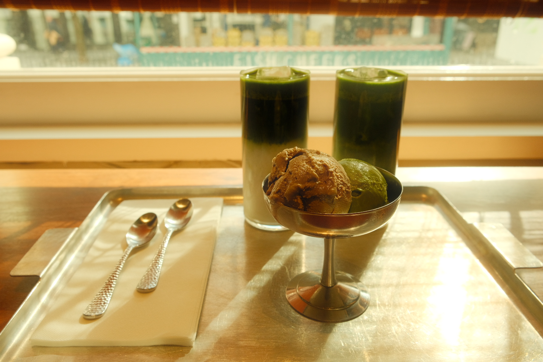 Iced matcha lattes and a silver coupe of chocolate and matcha gelato on a café tray by a sunlit window in New York.