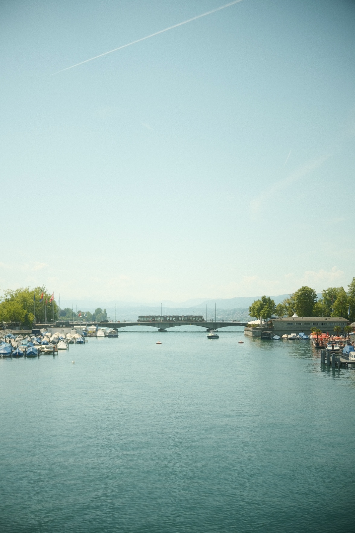 Zurich harbor view with moored boats and a bridge under a hazy blue sky.