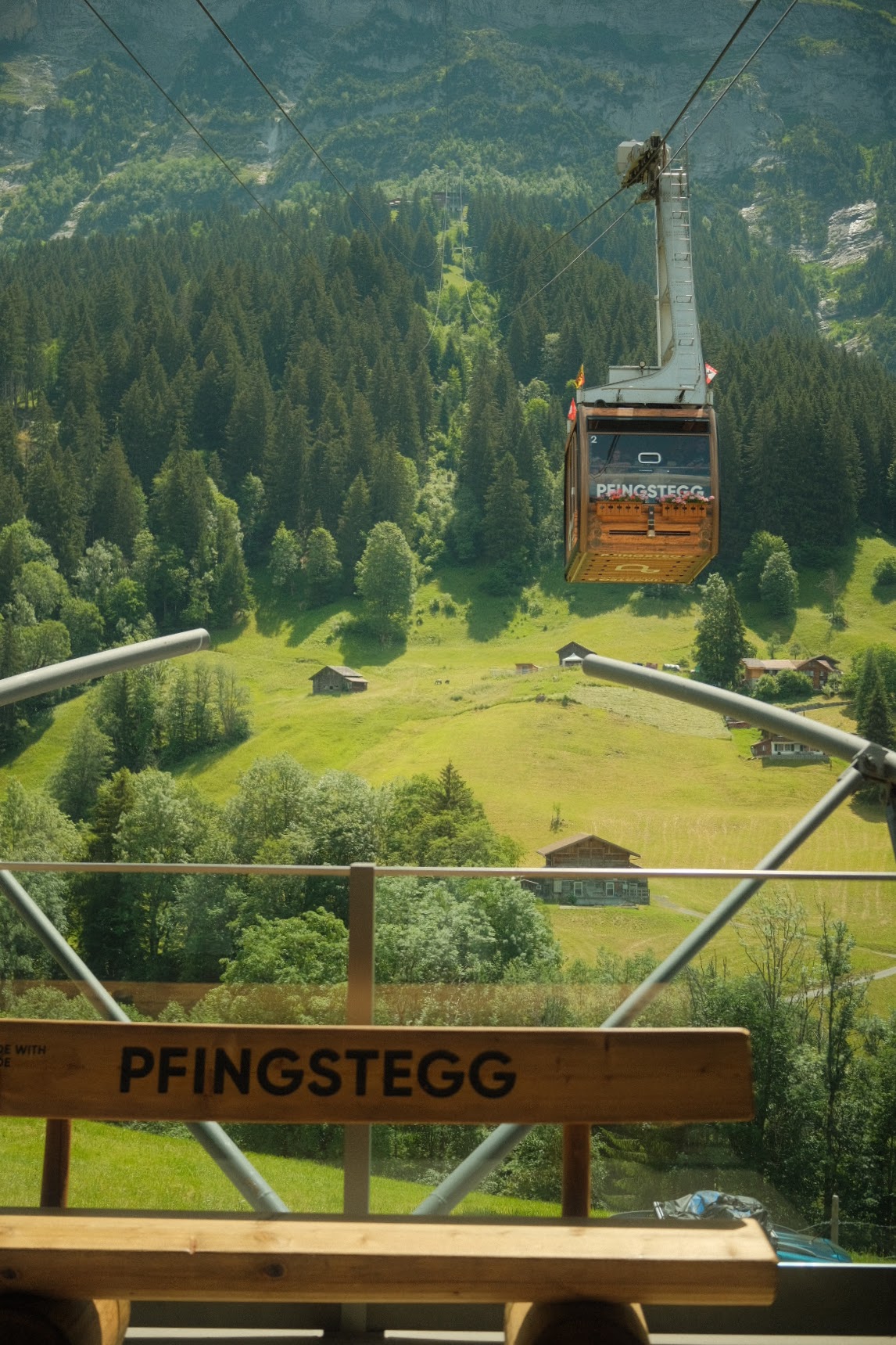 Pfingstegg gondola cresting the green Grindelwald valley, seen past a wooden Pfingstegg sign.