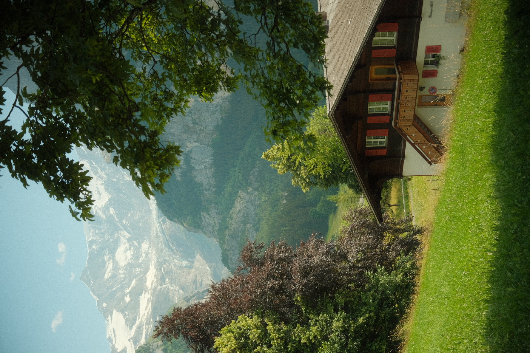 Traditional Swiss chalet with red shutters framed by trees, snow-dusted peaks rising behind.
