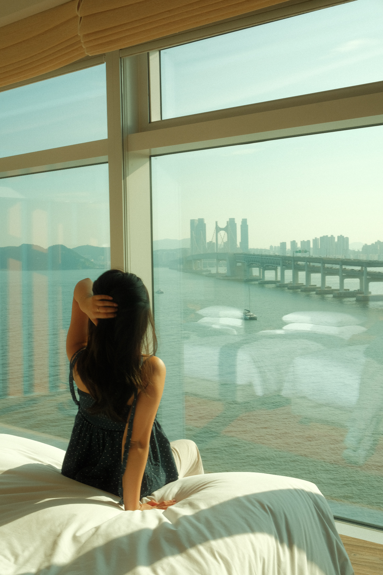A guest sitting on a hotel bed with a view of Busan and the Gwangan Bridge.
