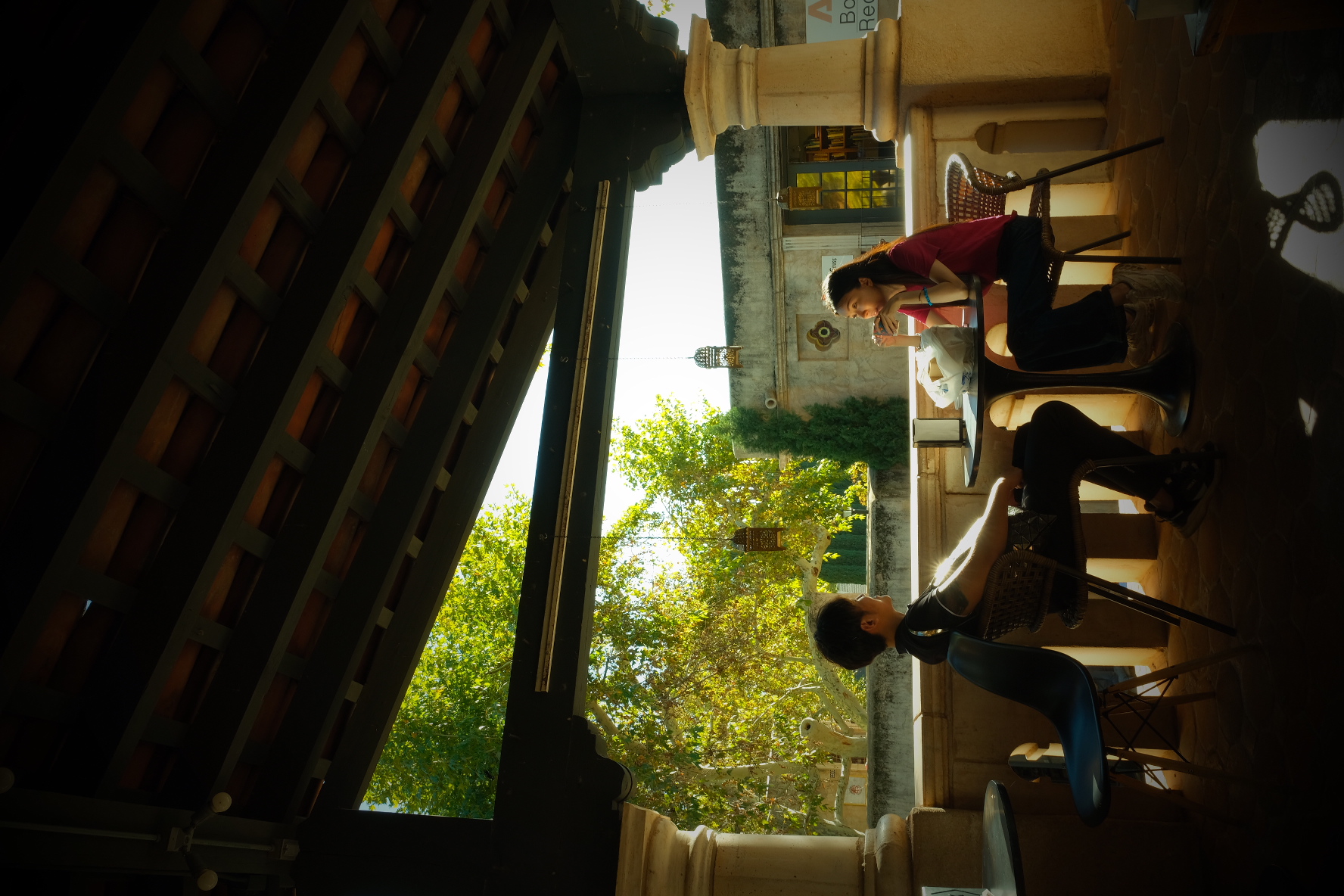 Young people at bistro tables beneath a wood-lattice roof on a sunlit café terrace in Arizona.