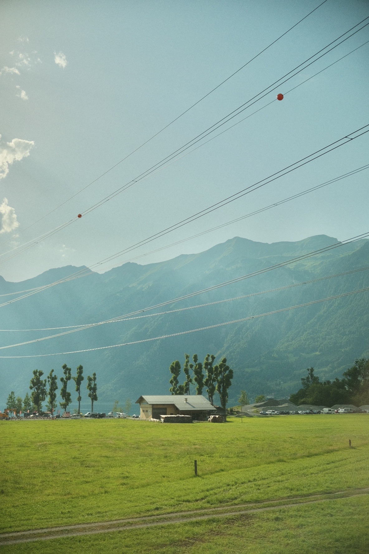 Lone barn in a green Alpine meadow beneath hazy mountains, power lines crossing the sky.